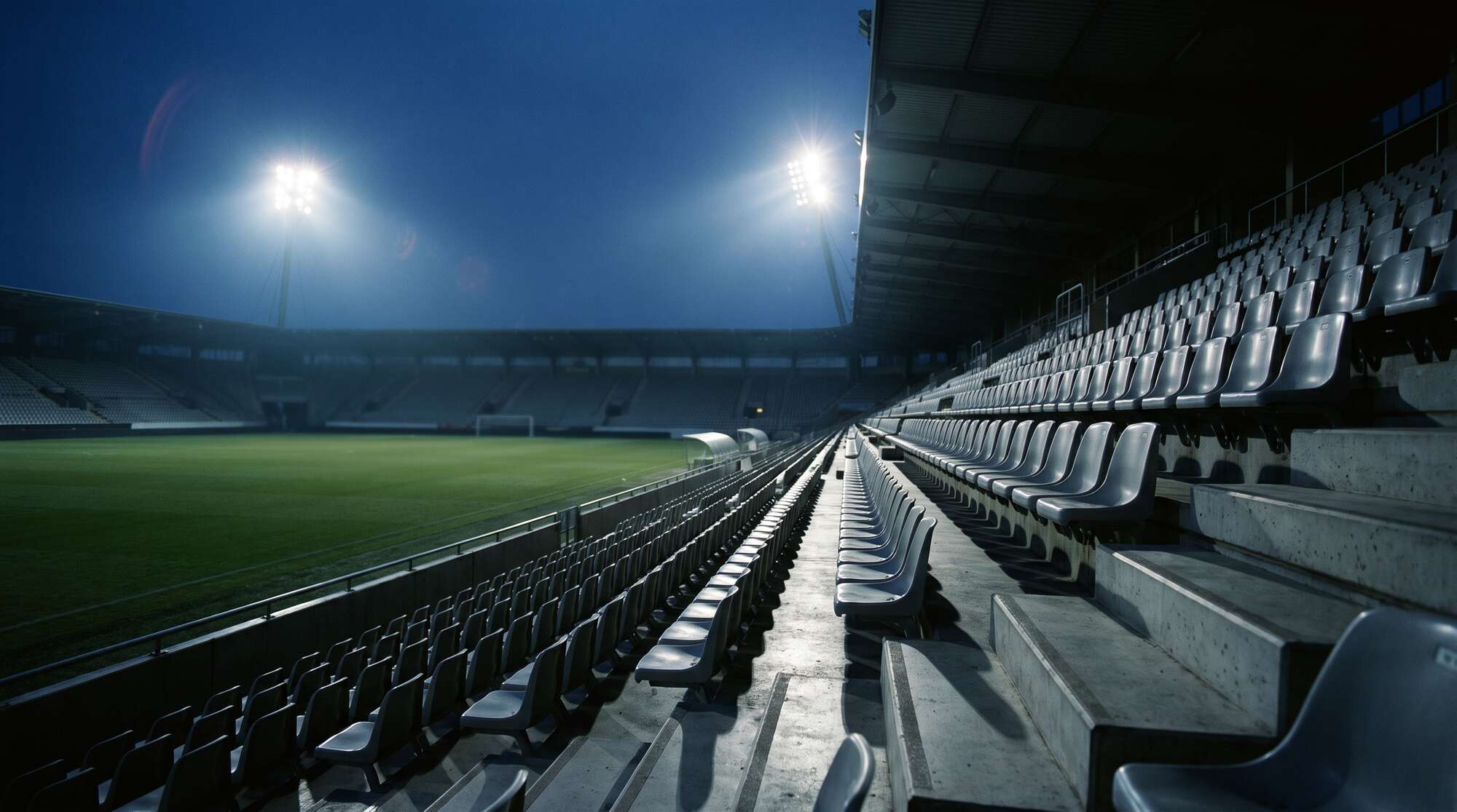 Floodlights coming on at dusk, view from the empty stand across the pitch
